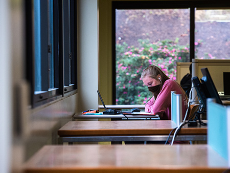 Student with mask on looking at laptop in a library. 