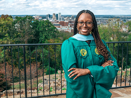 Nakia Coleman in her graduation cap and gown standing with arms crossed with Birmingham city skyline behind her. 