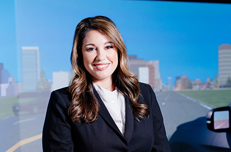 Environmental headshot of Dr. Despina Stavrinos, PhD (Associate Professor, Psychology) standing in front of the Driving Simulator in the TRIP (Translational Research for Injury Prevention) Laboratory, 2020.