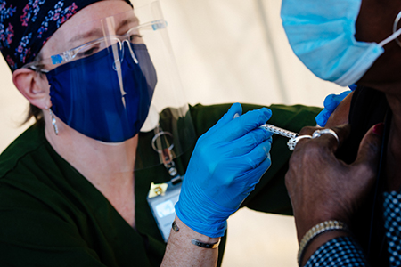 A female healthcare worker is wearing PPE (Personal Protective Equipment) head cover, face mask, face shield, and gloves while administering the COVID-19 vaccine to a black male at the UAB COVID-19 Vaccination Site at Parker High School on February 10, 2021/.