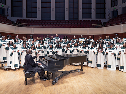 From side, Dr. Reginald Jackson, EdD (Instructor, Music; Director, UAB Gospel Choir) is playing a Steinway & Sons grand piano while directting students in UAB Gospel Choir wearing their robes while on stage in the Jemison Concert Hall at the Alys Stephens Performing Arts Center, 2019.