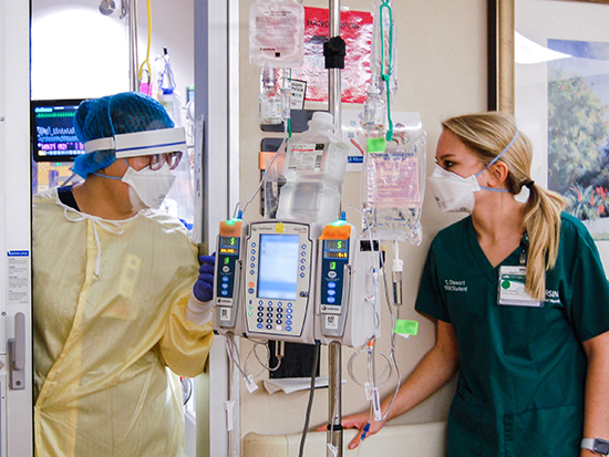 A female healthcare worker is wearing PPE (Personal Protective Equipment) head cover, face mask, gown, and gloves while talking to a female nursing student wearing scrubs and a PPE face mask on the floor with COVID-19 (Coronavirus Disease) patients, January 2021.