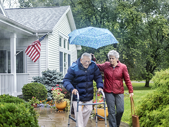 Looking through a raindrop splotched car window while waiting for a real person, real life, senior adult 95 year old elderly man wearing a parka-style winter coat. He is pushing his mobility walker down the home footpath ramp while walking side by side with his senior adult woman home caregiver daughter. She is holding an umbrella up above his head to protect him from the heavy rain storm. They are on their way out for some errands. He is a proud WWII military veteran who routinely displays the American flag - even sometimes (inadvertently) in the rain. Rochester, NY area suburban residential district. Selective focus beyond the people on the drenched flag and house.