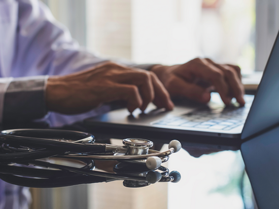 Male doctor in white coat typing on laptop computer keyboard with medical stethoscope on the desk in medical room at clinic or hospital.