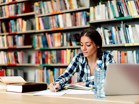 Female Student sitting in library searching information on laptop for her assignment. She is sitting at table surfing internet for notes for her study. Writing in her notepad.