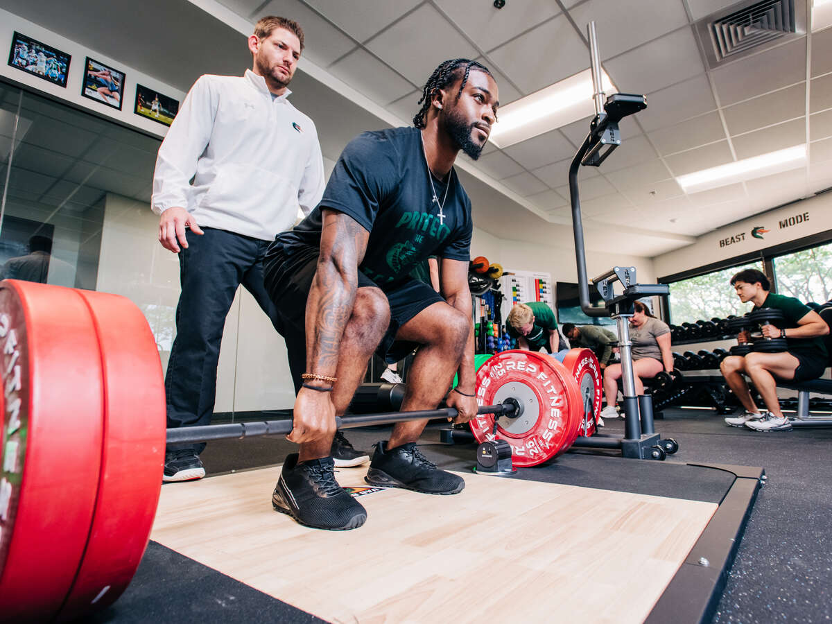 Student is lifting weights while faculty member stands behind him and spots. 