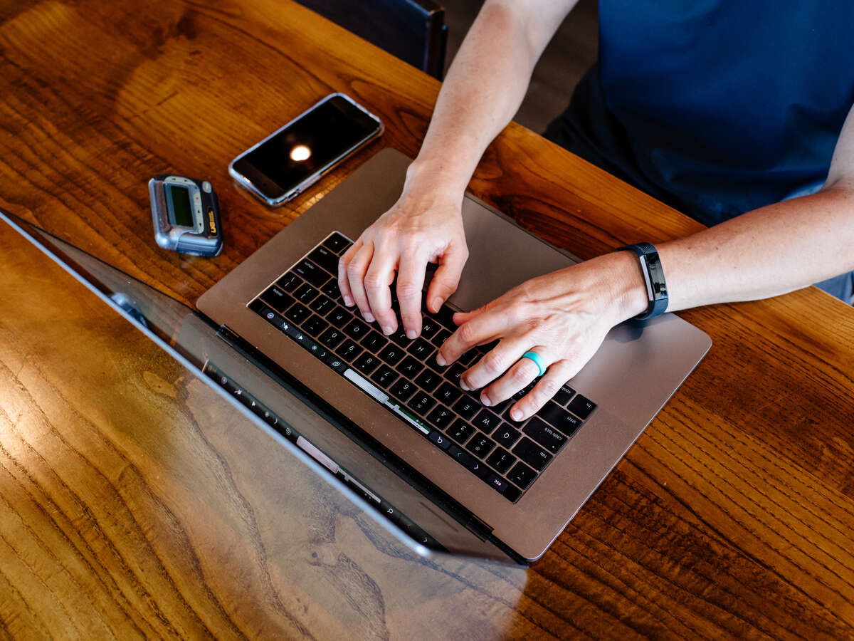 Person typing on a laptop at a wooden table, with a smartphone and fitness tracker nearby.