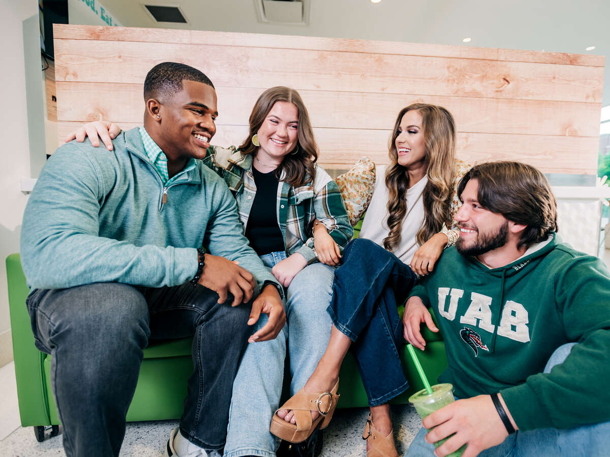 Collat School of Business. Four smiling students (one wearing a green UAB sweatshirt) are sitting on sofa in common space, April 2024.