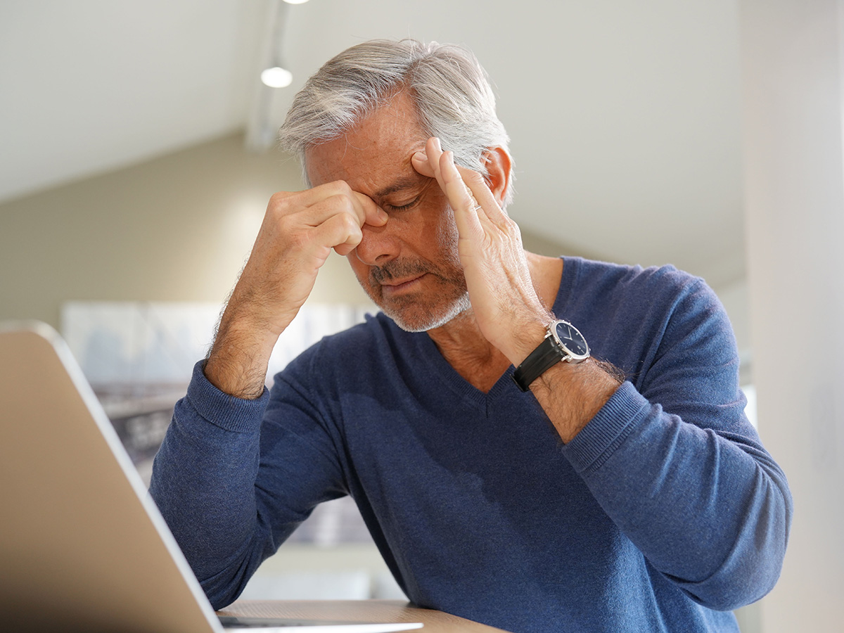 Gray haired man in blue shirt grabs nose with right hand and temple with left.
