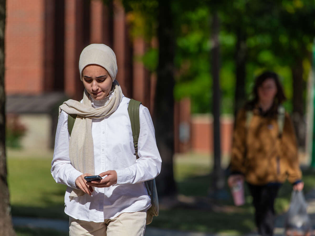 A student wearing a hijab and a backpack is looking at her mobile phone while walking on the Campus Green.