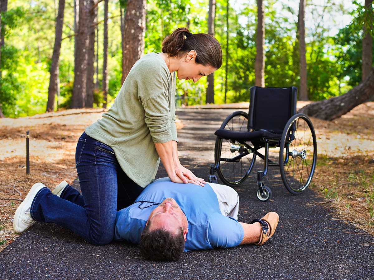 A woman kneels on a path outdoors and performs chest compressions on a man lying on their back, with a wheelchair nearby and trees surrounding the scene.