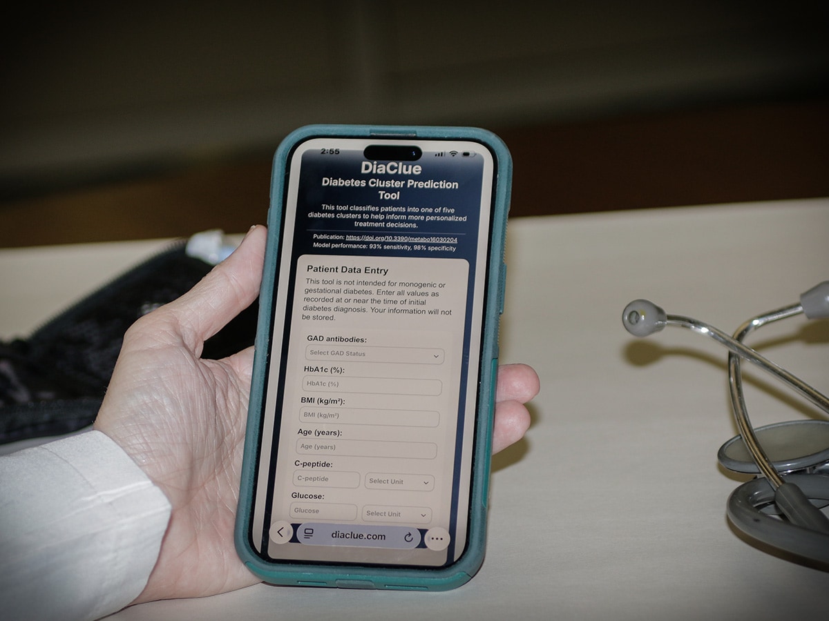 A hand holds a smartphone displaying the Diaclue webpage, with data entry fields visible on the screen. The phone rests on a light-colored surface beside a stethoscope, suggesting a clinical setting.