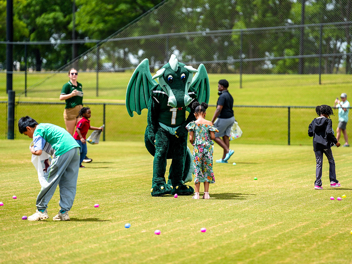 Outdoor image of children hunting bright, colored Easter eggs and a green Blaze the Dragon interaction with a child that is dressed in a floral outfit. 
