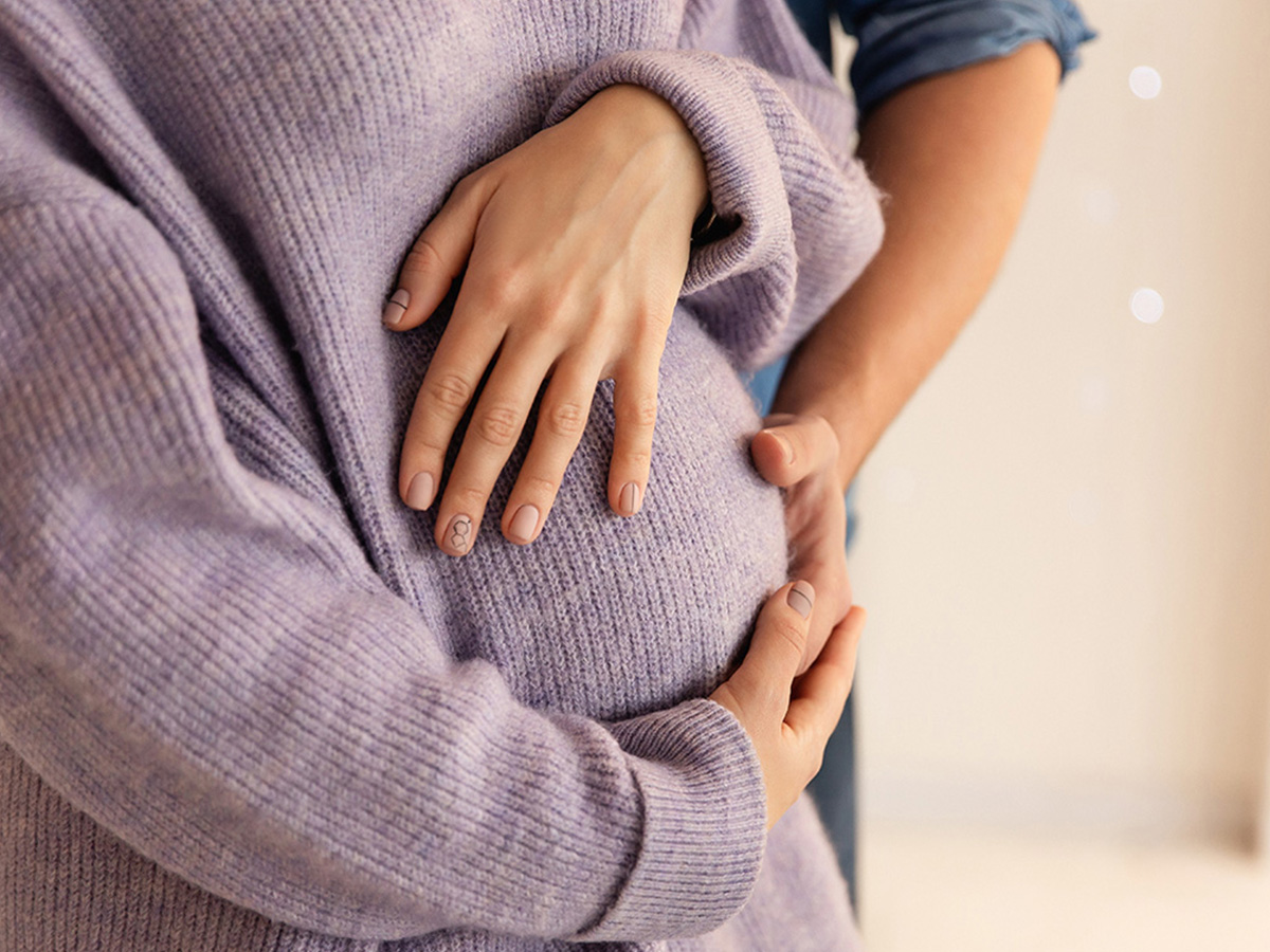 Close-up of hands resting on a women's rounded abdomen  wearing a lavender sweater, with another person’s arms gently wrapped around the waist from behind.
