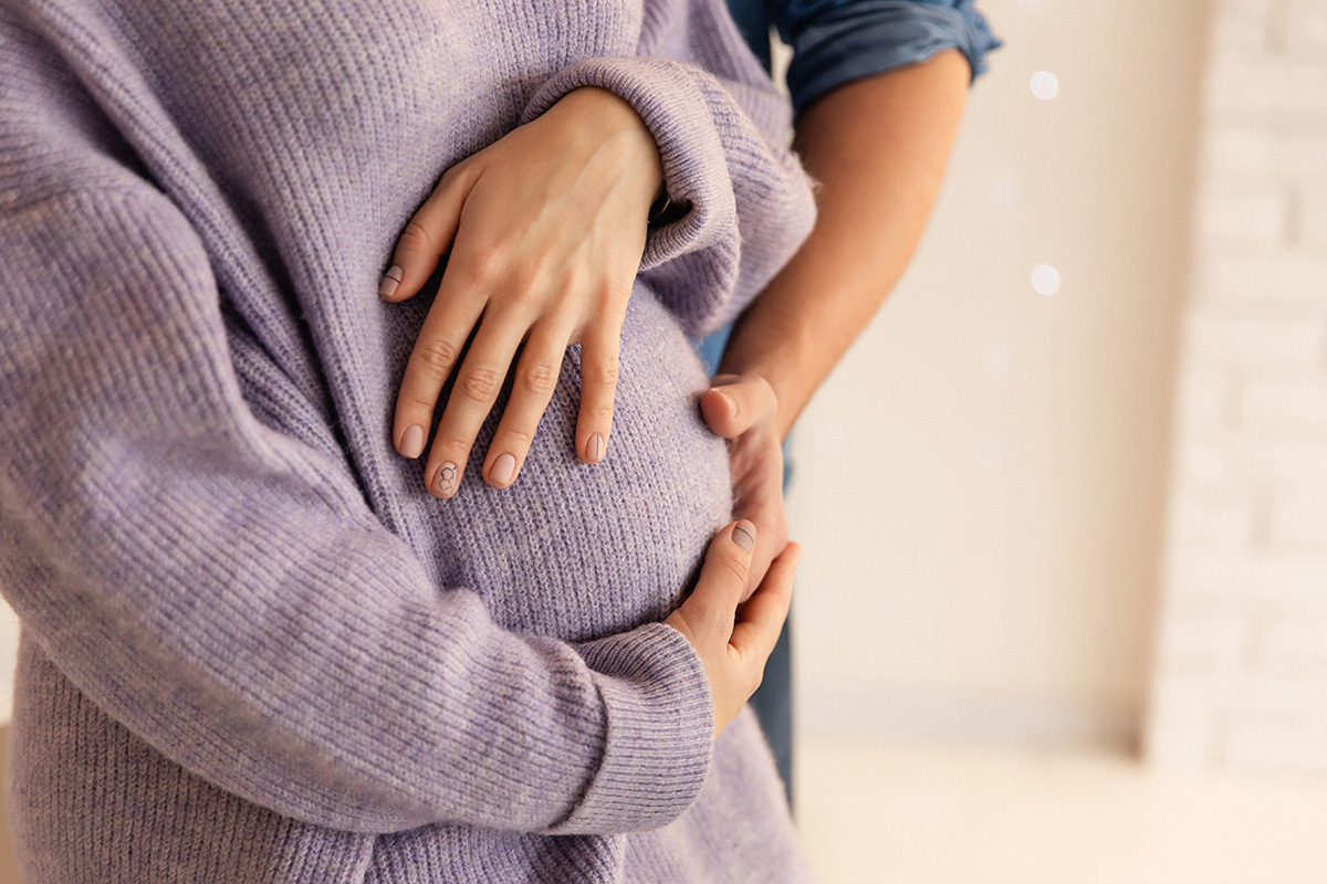 Close-up of hands resting on a women's rounded abdomen  wearing a lavender sweater, with another person’s arms gently wrapped around the waist from behind.
