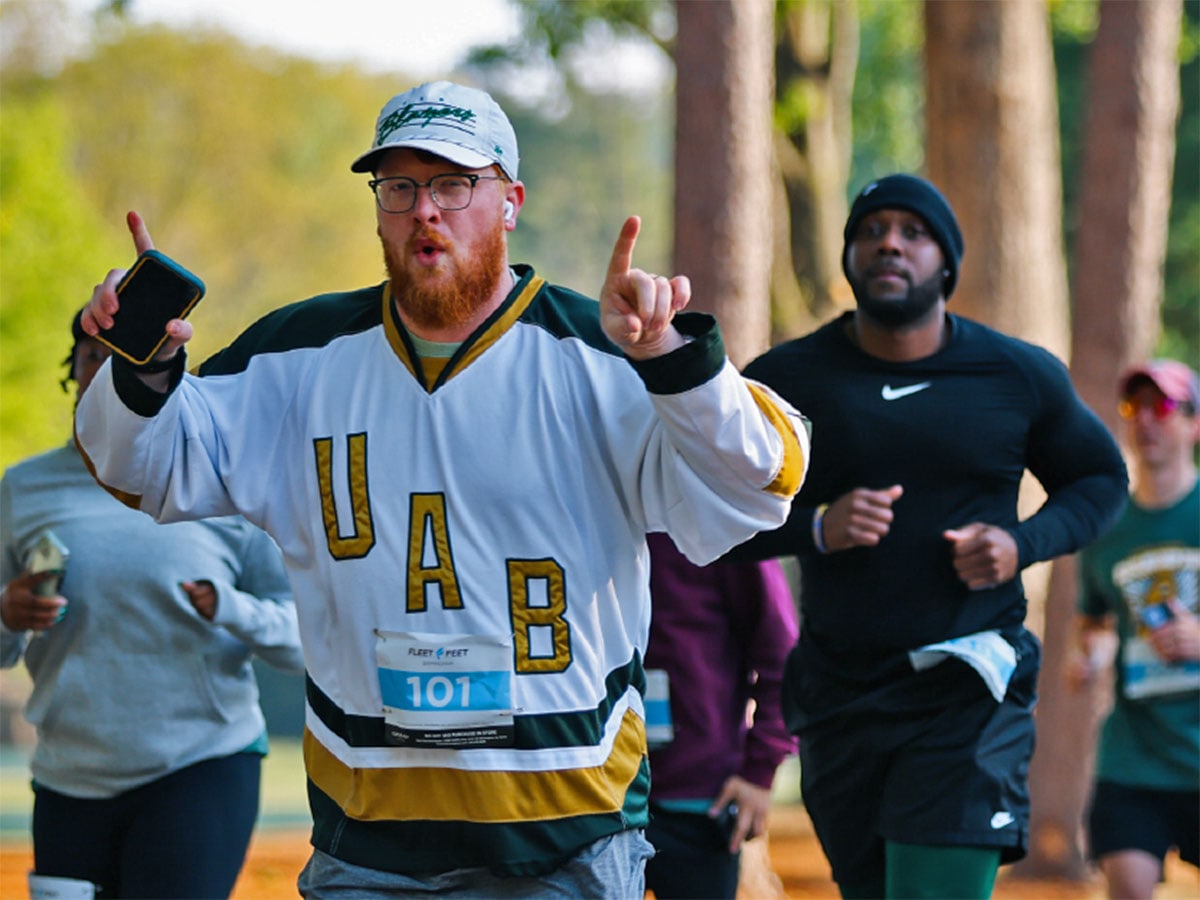 Participants run in the UAB Alumni Scholarship Race, including a man in a UAB jersey and hat making hand gestures in support of UAB.