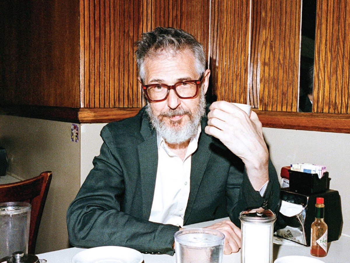 Ira Glass, a man with a beard and glasses wearing a dark suit jacket and white shirt, sits at a table in a restaurant holding a cup.