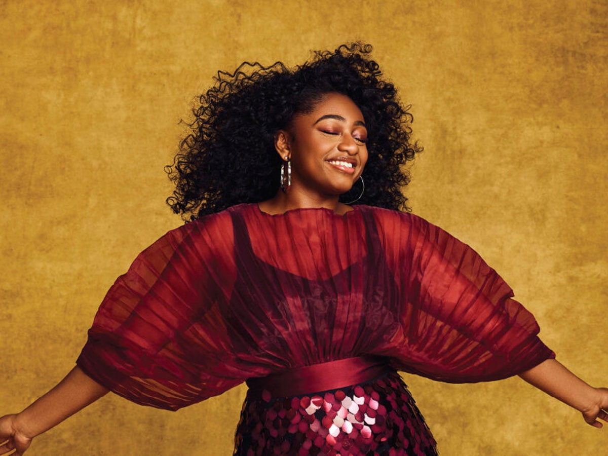 A woman wearing a red dress opens her arms wide and smiles with closed eyes, while standing in front of a gold wall. 