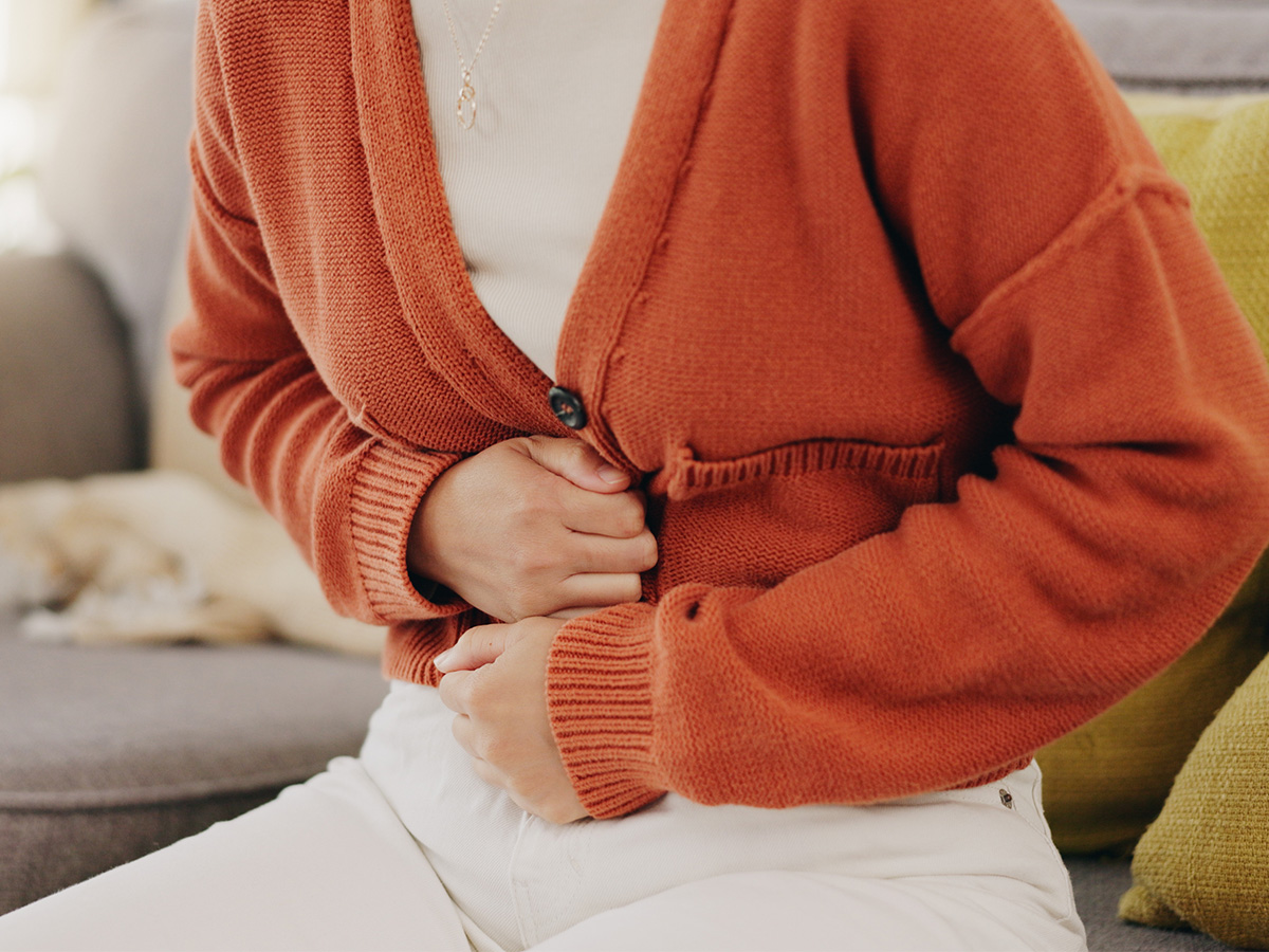 Body of a woman sitting on couch wearing an orange cardigan and white jeans with hands over her stomach.