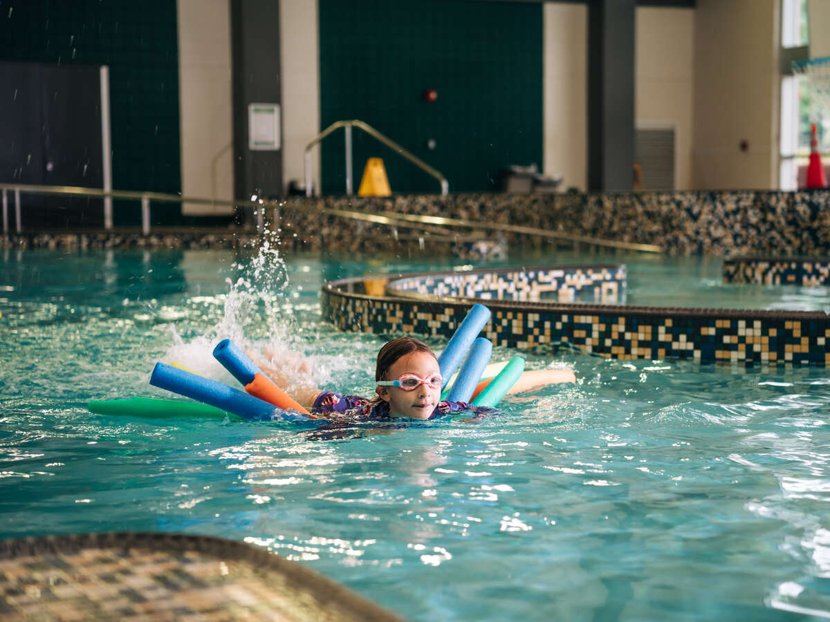 A girl is swimming with pool noodles in the indoor swimming pool at a URec summer camp in the University Recreation Center.