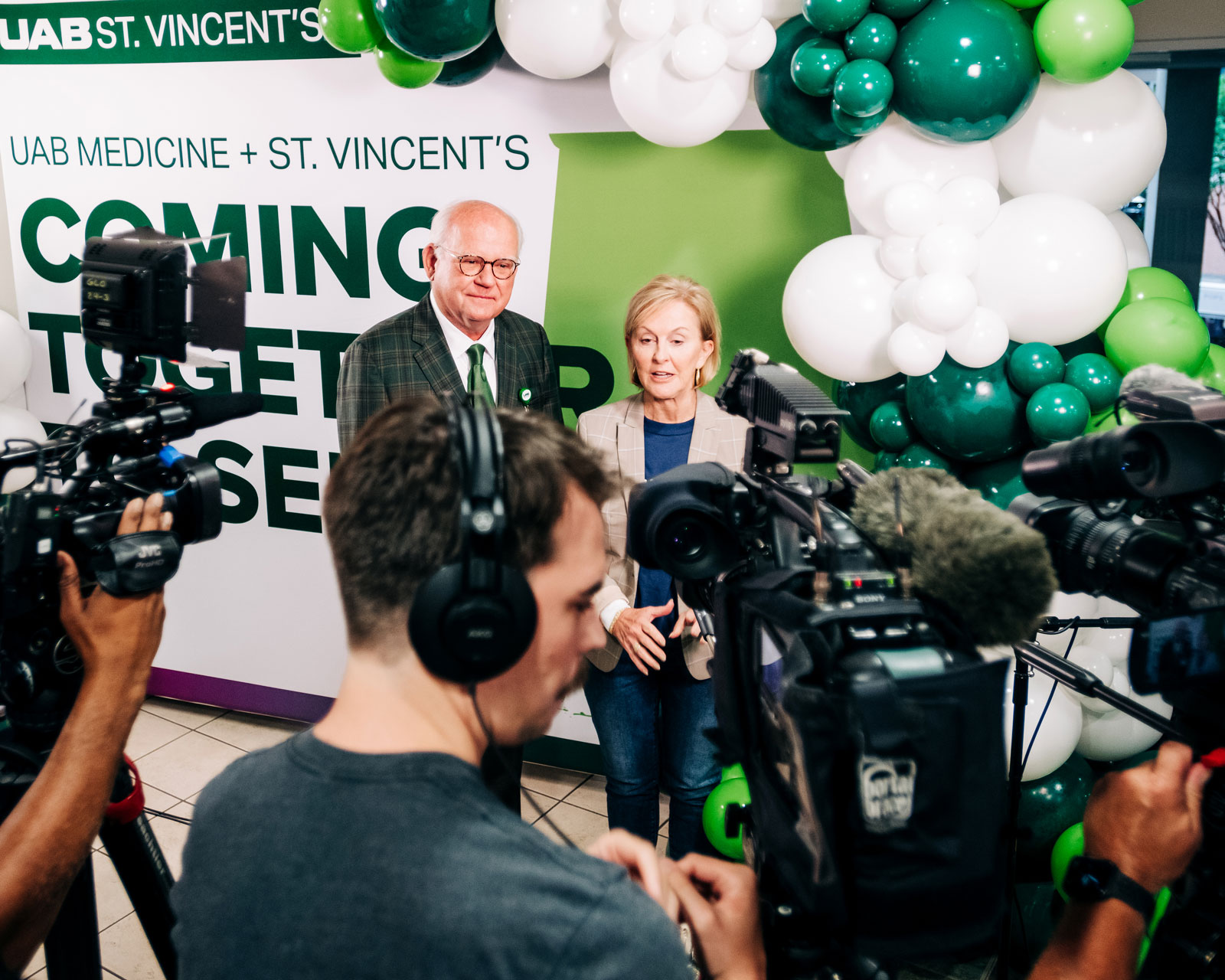 Two people stand in front of a sign reading 'Coming Together' surrounded by cameras and microphones. White and green balloons decorate the background.