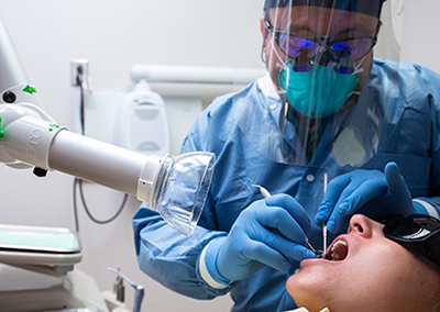 School of Dentistry. Dr. Augusto Robles, DDS (Associate Professor, General Dentistry) is wearing a PPE (Personal Protective Equipment) face shield, face mask, and gloves while performing a dental examination on a female patient who is sitting in a dental chair in front of the ExtraOral Vacuum Aspirator, which is a dental extraoral suction unit, used to capture and contain spit and other particles during dental procedures during COVID-19 (Coronavirus Disease) pandemic, 2020.