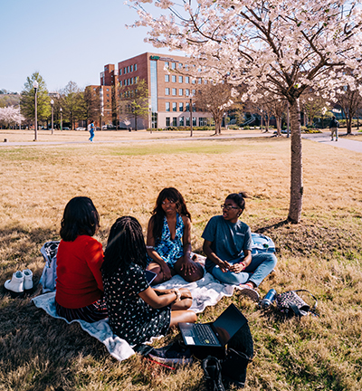 A group of ethnically diverse women, Ayesha Abuzer (Student, Pre-Immunology), Alexandria Martin (Student), Faith Williams (Student, Digital Forensics), and Joline Ugbo (Patient Care Tech, Nursing Services - Highlands) are sitting on a blanket on the grass in front of Blazer Hall and Commons on the Green on the Campus Green, Spring 2022.