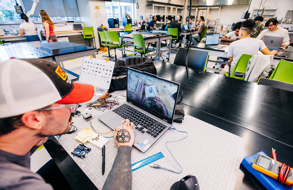A wide-angle view of the Design x Prototyping Lab at the UAB School of Engineering with students at work at each of several visible tables.