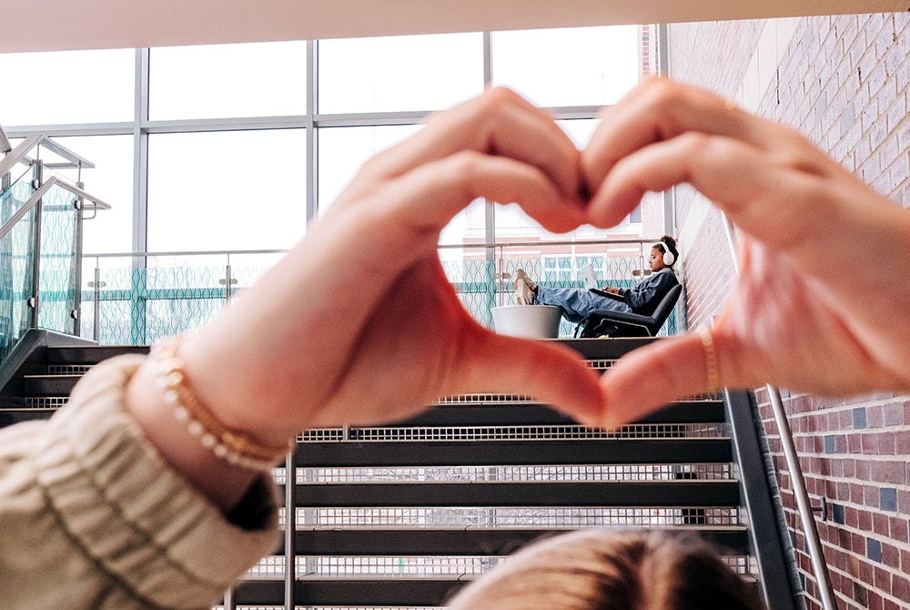 In the foreground, a figure is making a heart with her hands connected and the heart frames a student, wearing headphones, studying on the landing of the stairs at the Hill Center