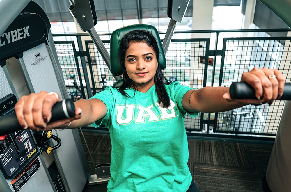 A student wearing a green U A B shirt looks into the camera while at full extension on the chest press machine at the Rec Center