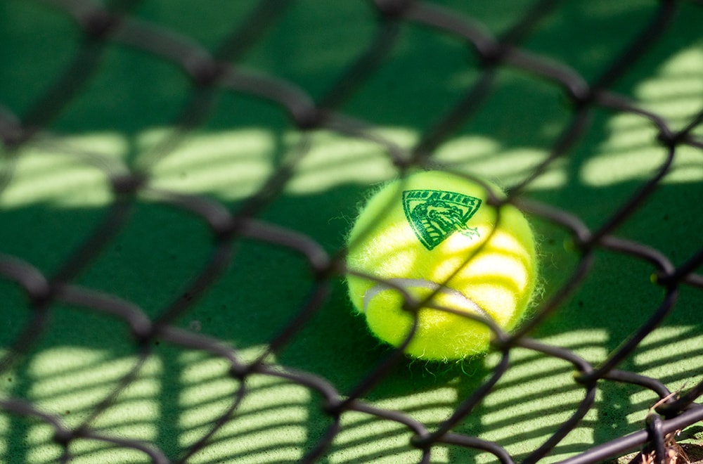 A tennis ball with UAB Blazers logo sits in shadows cast on the court by the net