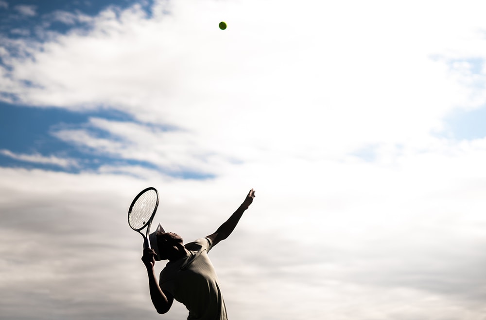 A tennis ball is at the height of its arc while a waiting Men's Tennis player is poised to strike it with both framed against a cloudy sky