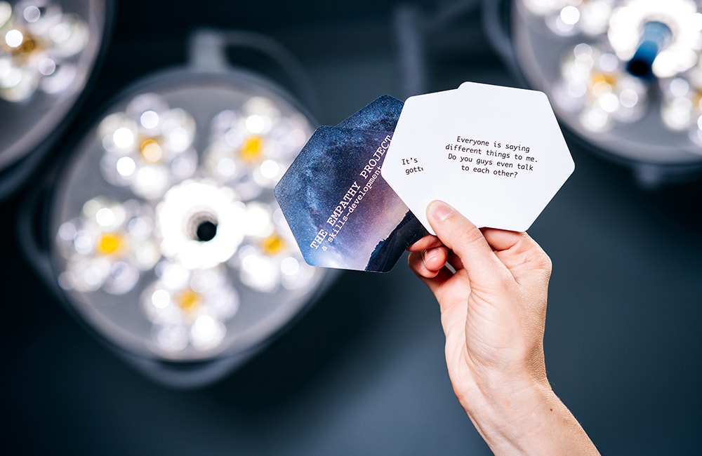 A hand holds four cards from The Empathy Game up to a light. The topmost card reads, "Everyone is saying different things to me. Do you guys even talk to each other?" 