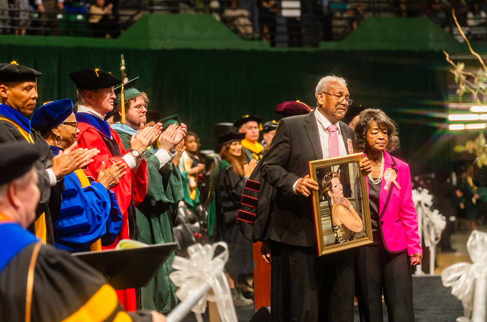 A man in dark suit and pink tie and a woman in pink suit jacket hold a portrait of a young woman while UAB faculty, students and leaders on the podium stand and applaud