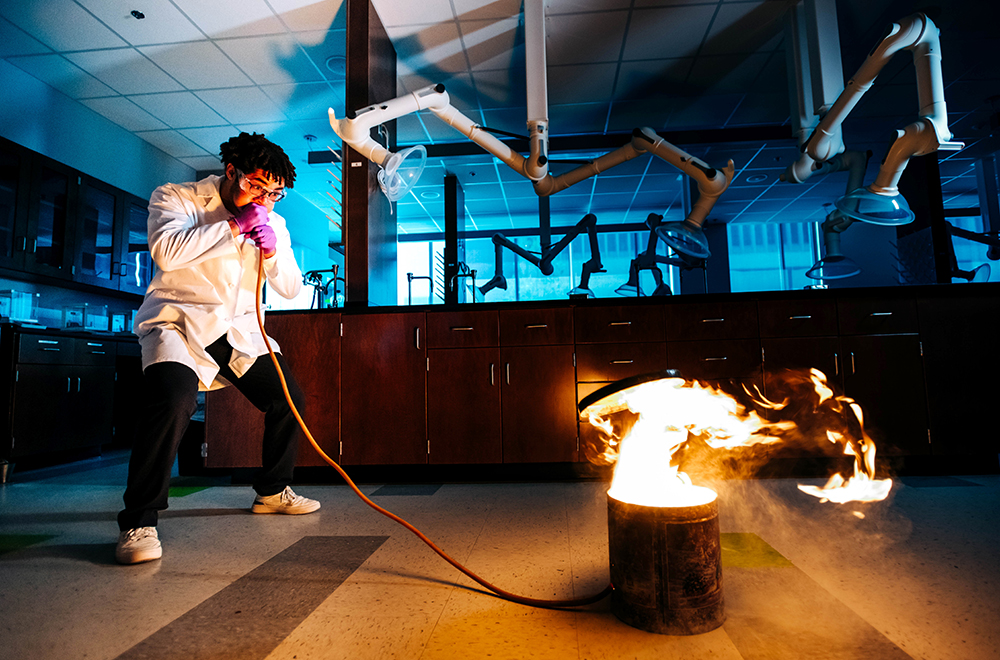 In a UAB chemistry lab, a white-coated student blows into a orange tube connected to a canister on the ground, while flames burst from the canister