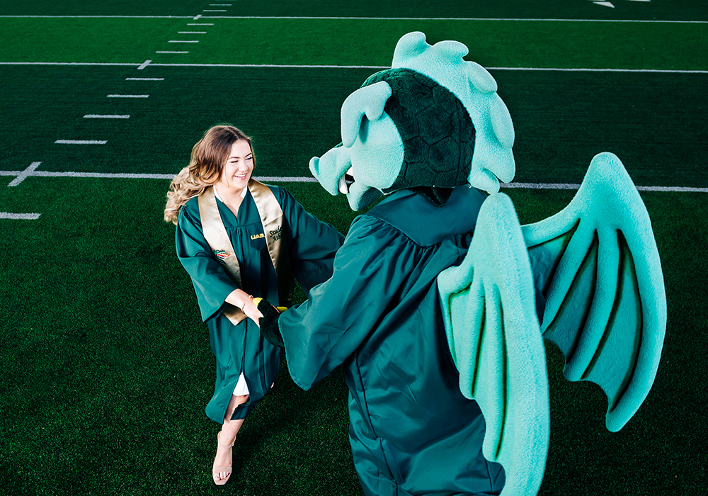 U A B mascot Blaze the dragon holds hands in a dance with a smiling student wearing a graduation gown and gold sash on the field at the U A B Football Practice Facility