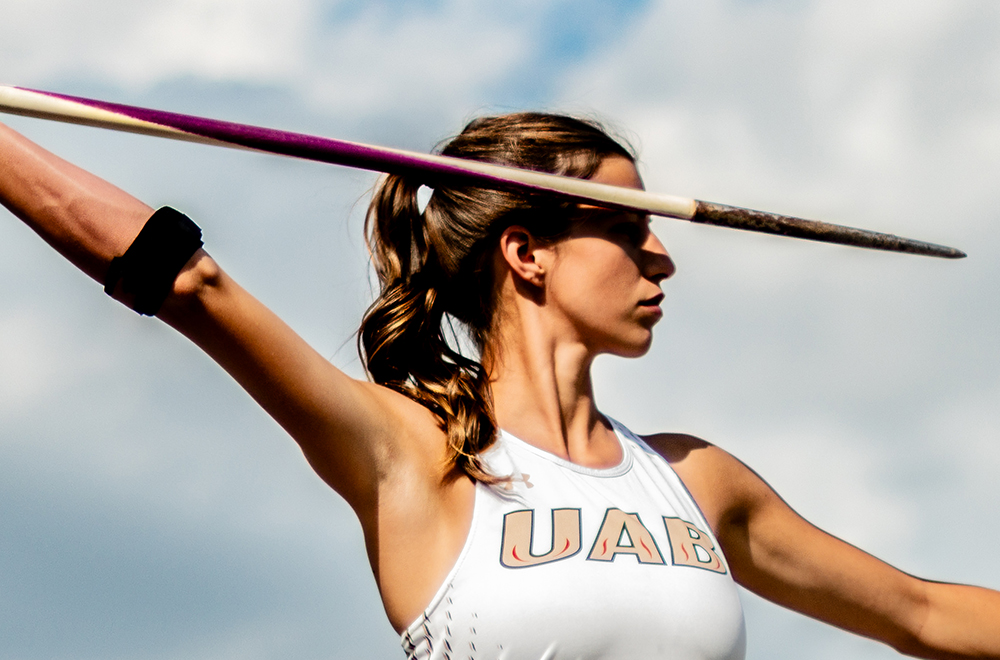 A female track athlete, seen from the waist up, holds a javelin poised in her right hand, with the shadow cast by the sun over her eyes