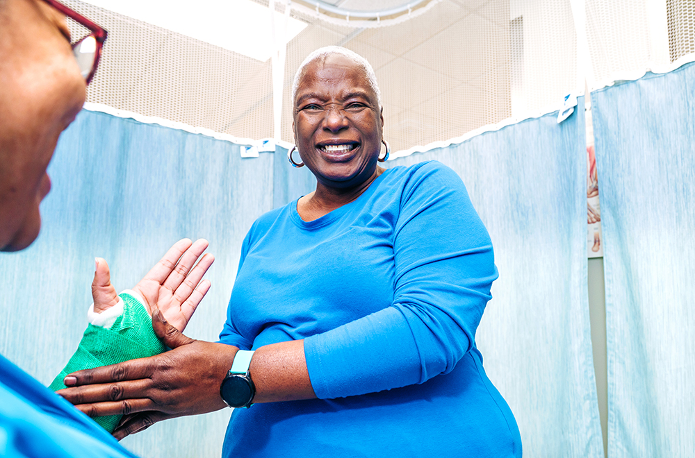 A smiling woman in blue scrubs looks at the camera while applying a green wrap to the arm of a seated model in a U A B clinic