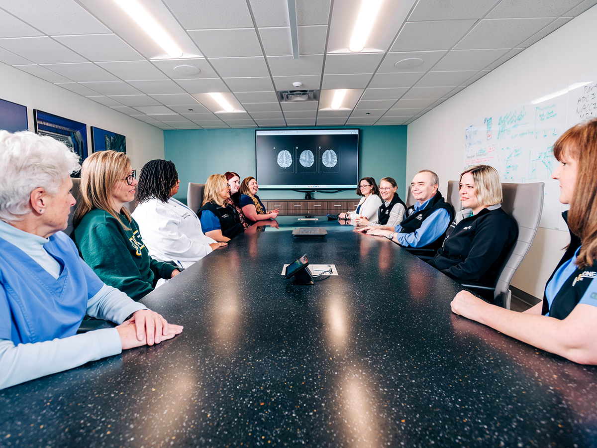 Members of the care team at the UAB Brain Aging and Memory Clinic gather around a conference table, with a screen showing mock brain scans behind them