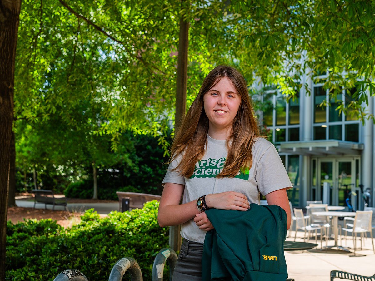 Amelia Hawkins, with her green graduation gown draped over her arm, stands under the trees outside Heritage Hall
