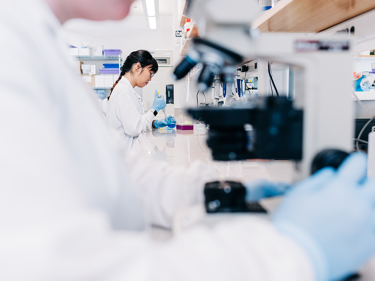 A researcher in a UAB lab is in focus in the background, pipetting into a petri dish, while an out of focus lab member in the foreground looks into a microscope with only her chin visible
