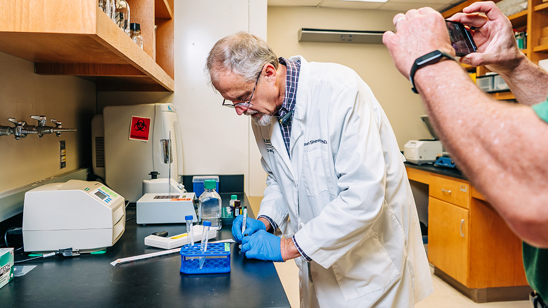 Dr Cameron Sheeler, wearing a lab coat, works on a lab test while Kevin Storr films with his phone