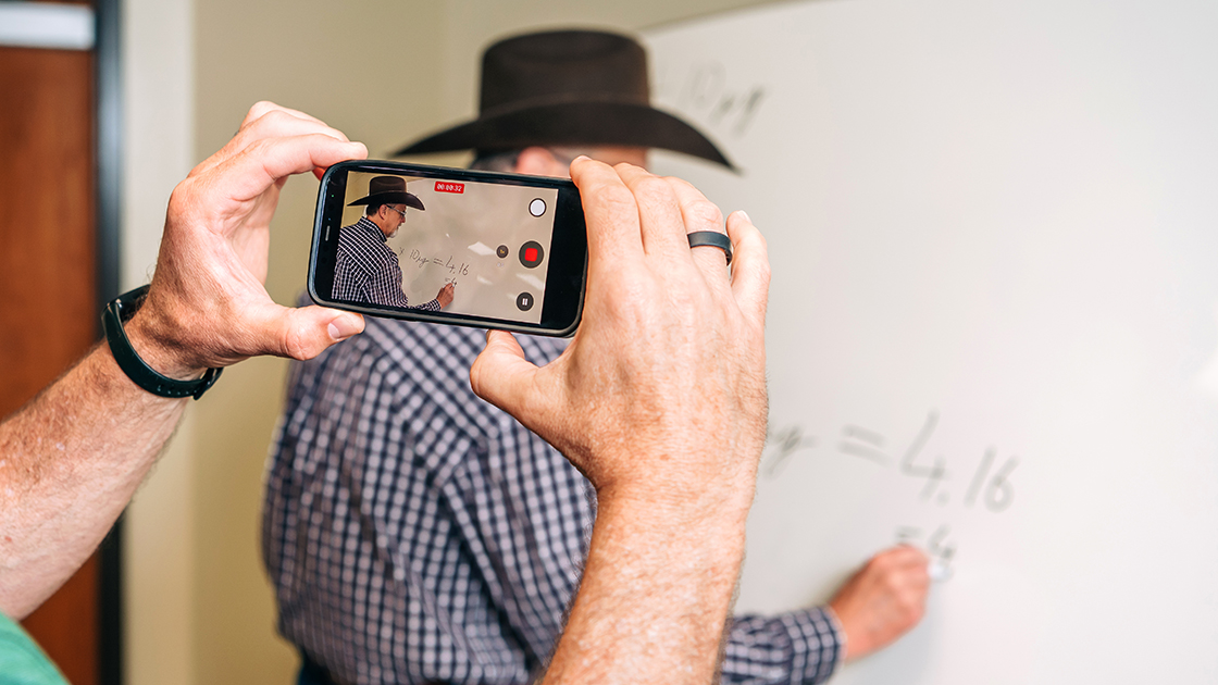 Dr. Sheeler, wearing a cowboy hat and writing on a whiteboard, is seen in focus on the screen of Kevin Storr's phone while Sheeler is also partly visible blurred behind