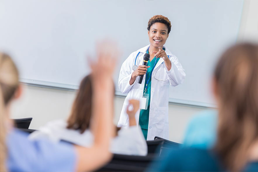 Woman in front of a whiteboard, wearing a medical white coat, points to someone in the foreground with a raised hand. 