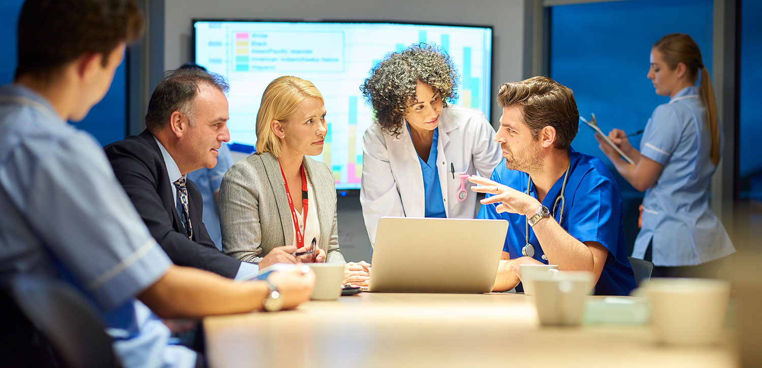 Group of people gathered around laptop talking with a chart on a screen in the background.