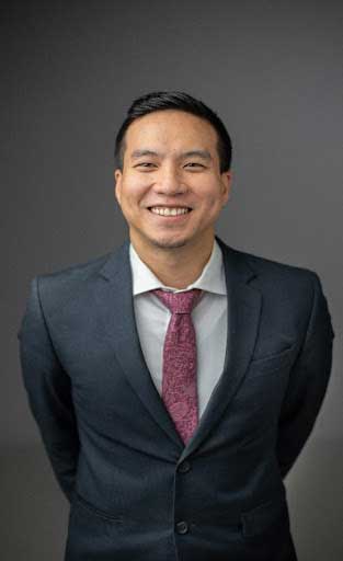 Dr. Byron Lai smiles at the camera, holding his hands behind him. He has short dark hair and wears a dark suit, white collard shirt, and burgundy tie. 