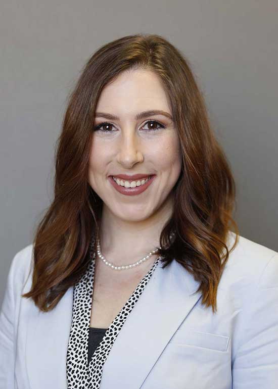 Dr. Ferguson has long red-brown hair and is wearing a white blazer over a black-and-white dotted shirt and a pearl necklace. 