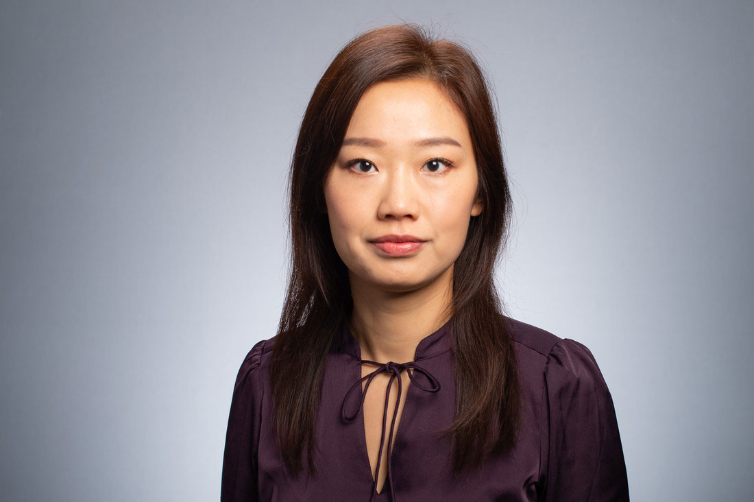 Dr. Jia Li has long dark brown hair. She's wearing a dark burgundy blouse with a ribbon tie at the neck. 