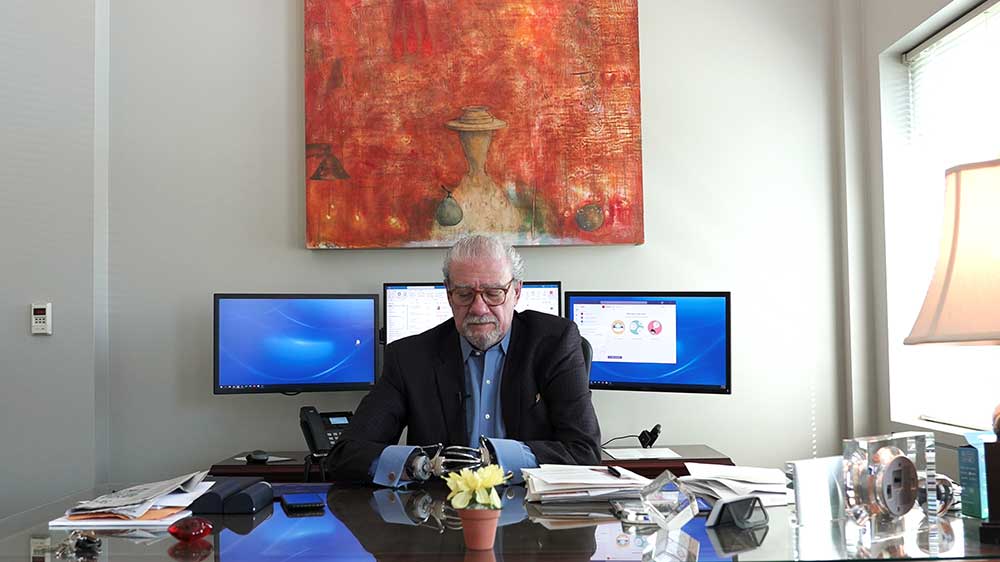 Kemp at office desk, three monitors behind him, an orange painting on the wall. 