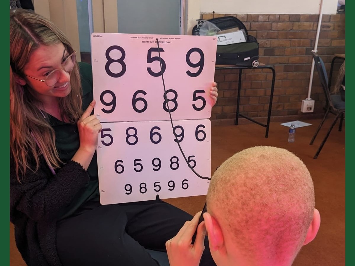 Smiling UAB OT student holds a numbered eye chart for a South African child to read.